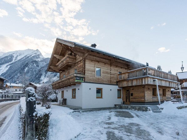 Un chalet en bois doté d’un balcon, une enseigne indiquant « HOLZ AU » et un paysage montagneux en arrière-plan.