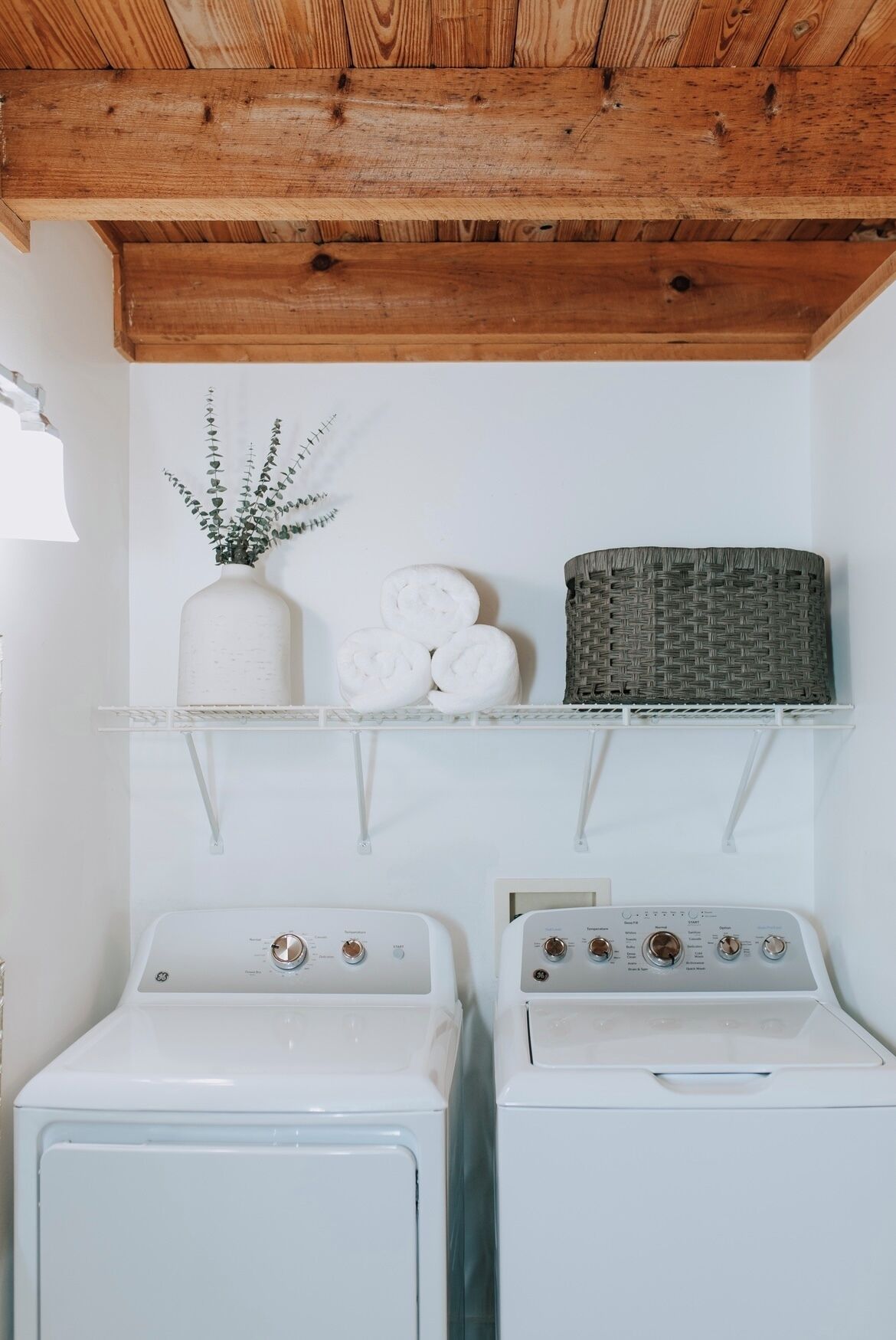 A laundry area with two washing machines, a shelf with a vase and rolled towels, and a woven basket.