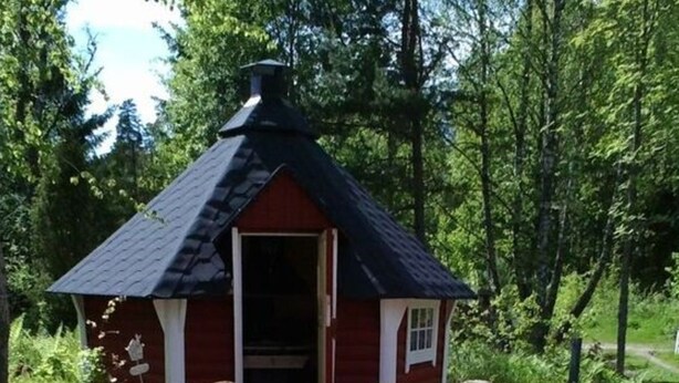 Uma pequena cabana de madeira com telhado escuro, porta e janela. Há uma rede em um deck de madeira em frente à cabana.
