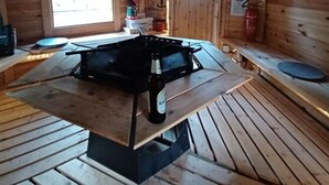 Interior de uma cabana de madeira, com uma lareira a lenha no centro, uma janela e piso de madeira.