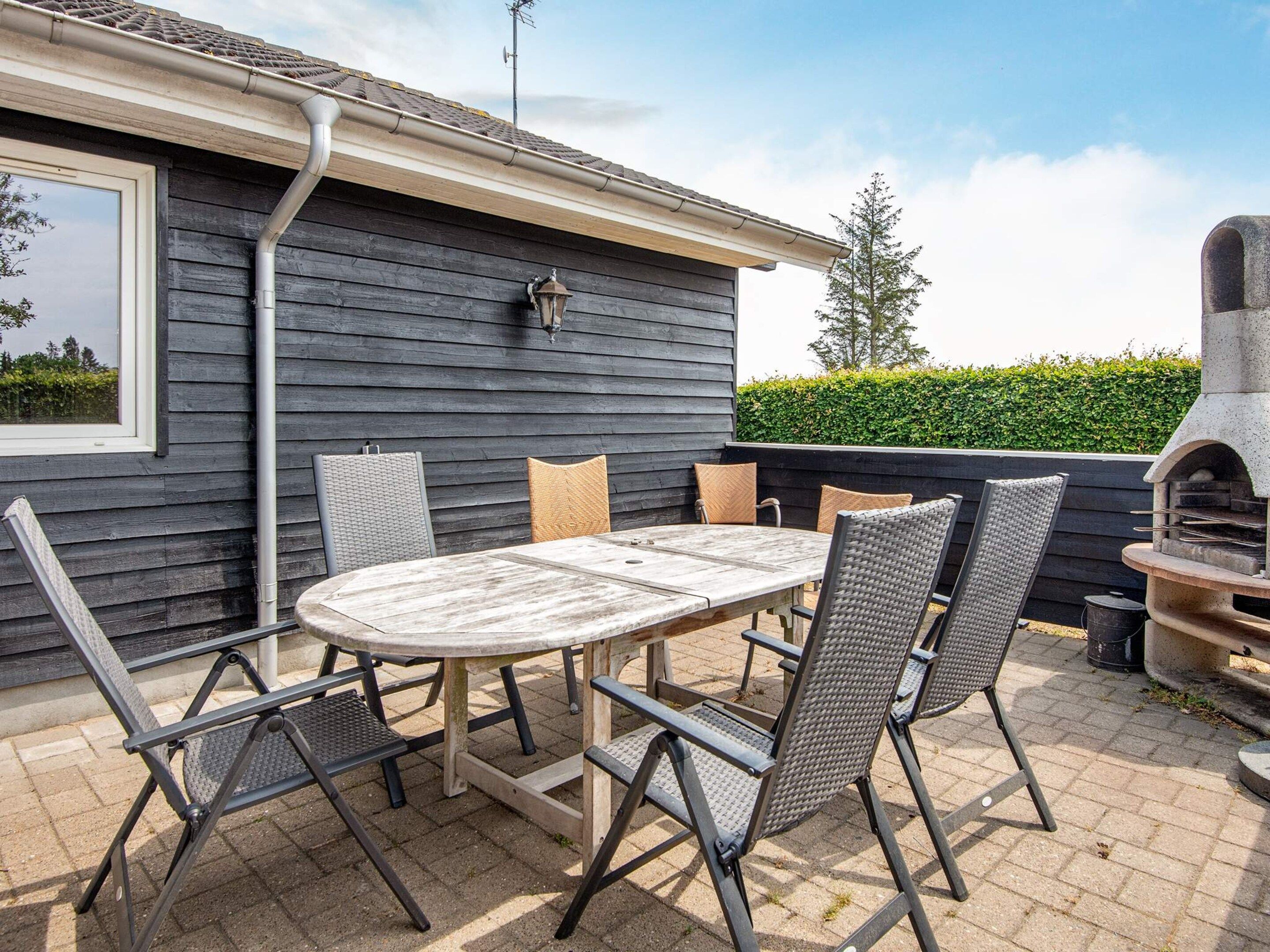 An outdoor patio with a round table and chairs, a grill, and a view of a hedge and trees.