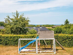 Un terrain de jeux avec un toboggan et une petite structure en bois, entouré de haies et d’arbres, avec des maisons en arrière-plan.