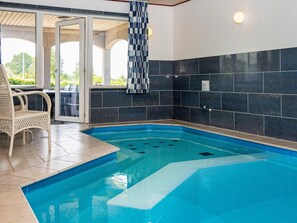 An indoor swimming pool with a tiled wall, a wicker chair, and a glass door leading to an outdoor area. - 4 Star Holiday Home in Glesborg (Glesborg)