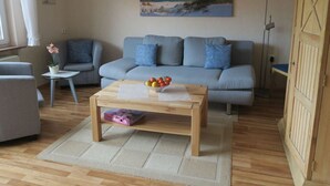 A living room with a grey sofa, a wooden coffee table with a fruit bowl, a small side table with a plant, and a painting of a beach scene on the wall. - North Sea House Schillig (Wangerland)