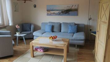A living room with a grey sofa, a wooden coffee table with a fruit bowl, a small side table with a plant, and a painting of a beach scene on the wall.