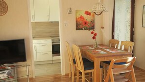 A dining area with a wooden table, chairs, and a vase of flowers. There is a television on a glass shelf and a kitchen in the background. - North Sea House Schillig (Wangerland)