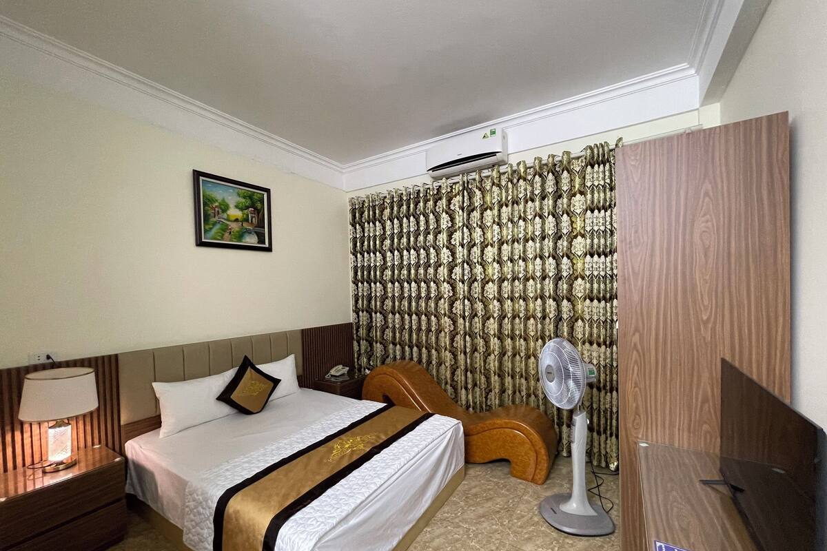A hotel room with a bed, a nightstand, a lamp, a fan, a wooden chair, and a wardrobe.