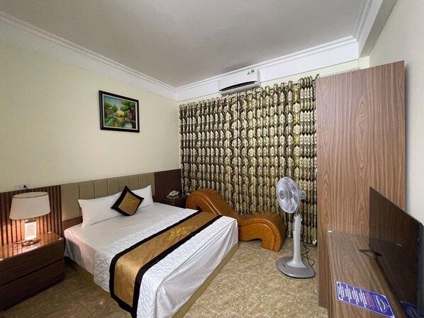 Una habitación de hotel con cama, mesita de noche, lámpara, ventilador, silla de madera y armario.