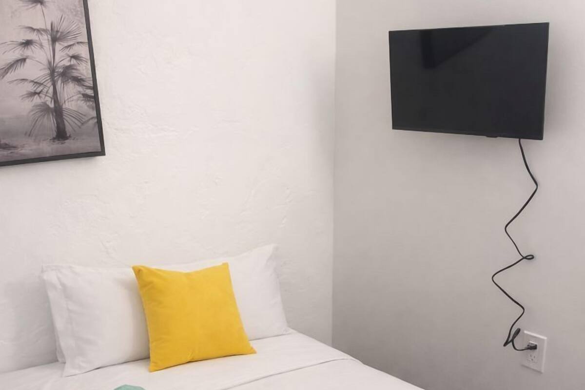 A neatly made bed with white and yellow pillows, a rolled-up towel, and a small gray pillow. A flat-screen TV is mounted on the wall above the bed.