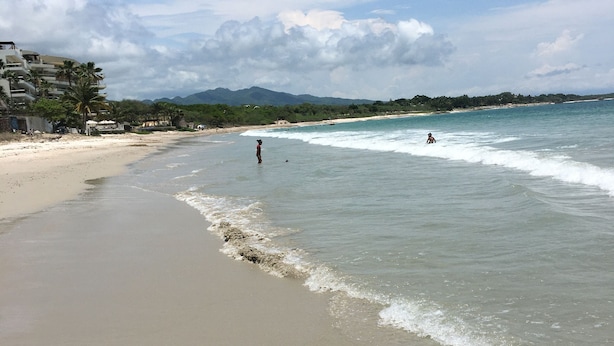 Une plage avec un ciel dégagé, des vagues douces et une chaîne de montagnes qui se dessine à l’horizon.