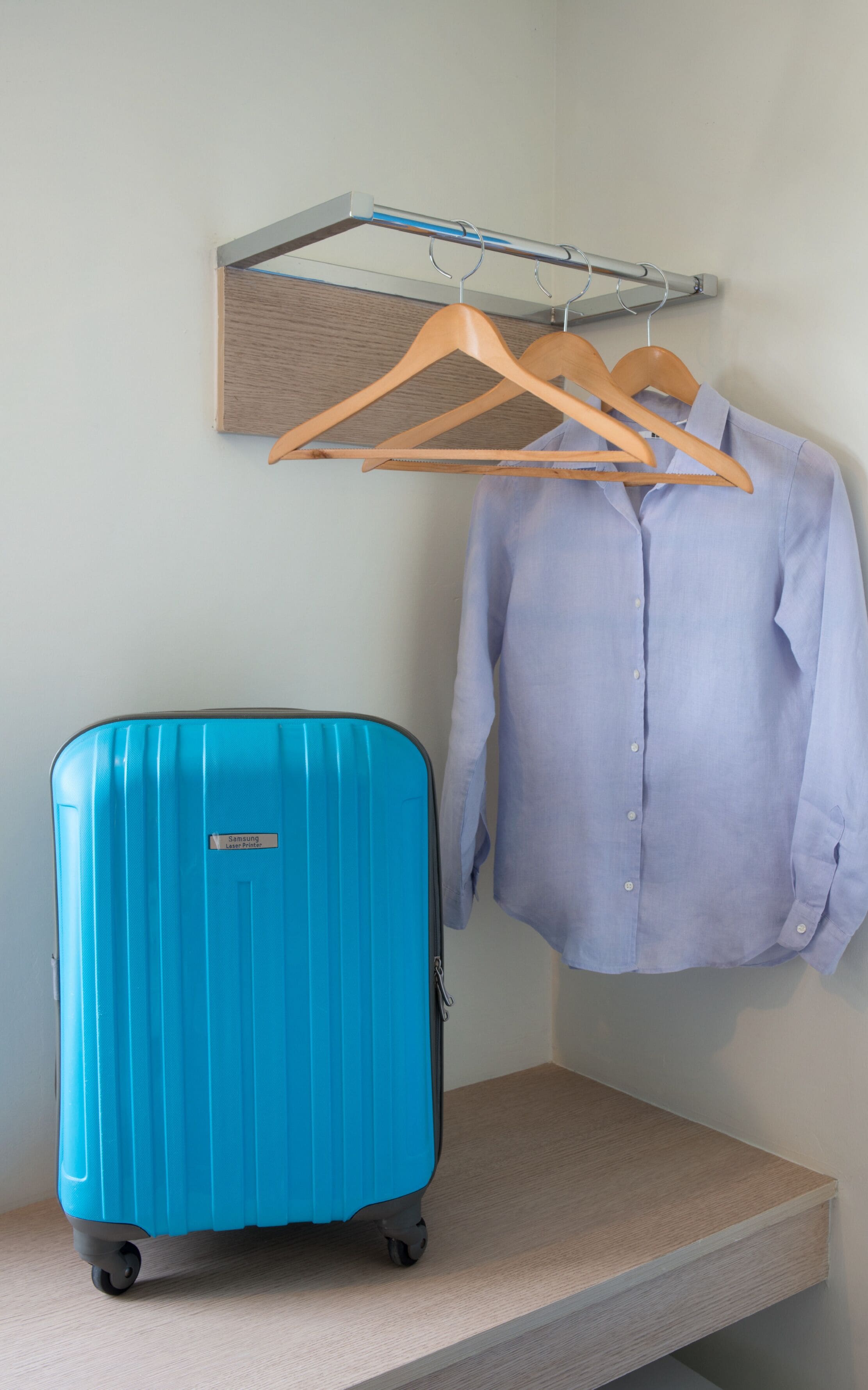 Una habitación con una maleta azul sobre una plataforma de madera, un perchero de madera con ropa y una pequeña estantería.