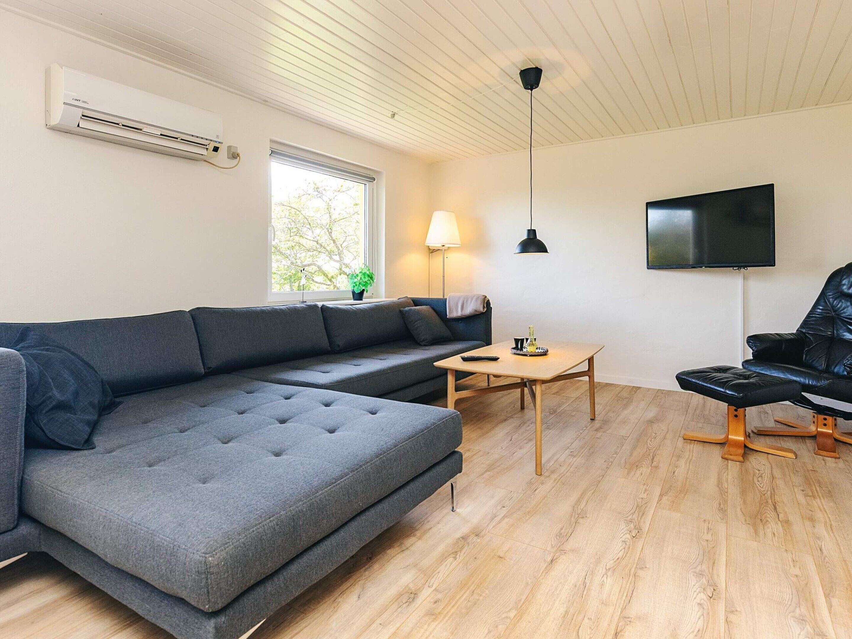 Una sala de estar moderna con un sofá modular, una pequeña mesa de madera, un sillón de cuero negro, un televisor montado en la pared y un equipo de aire acondicionado.