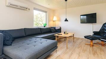 Una sala de estar moderna con un sofá modular, una pequeña mesa de madera, un sillón de cuero negro, un televisor montado en la pared y un equipo de aire acondicionado.