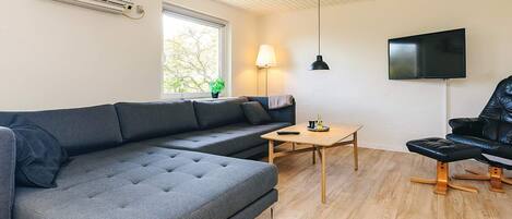 A modern living room with a sectional sofa, a small wooden table, a black leather chair, a TV mounted on the wall, and an air conditioning unit.