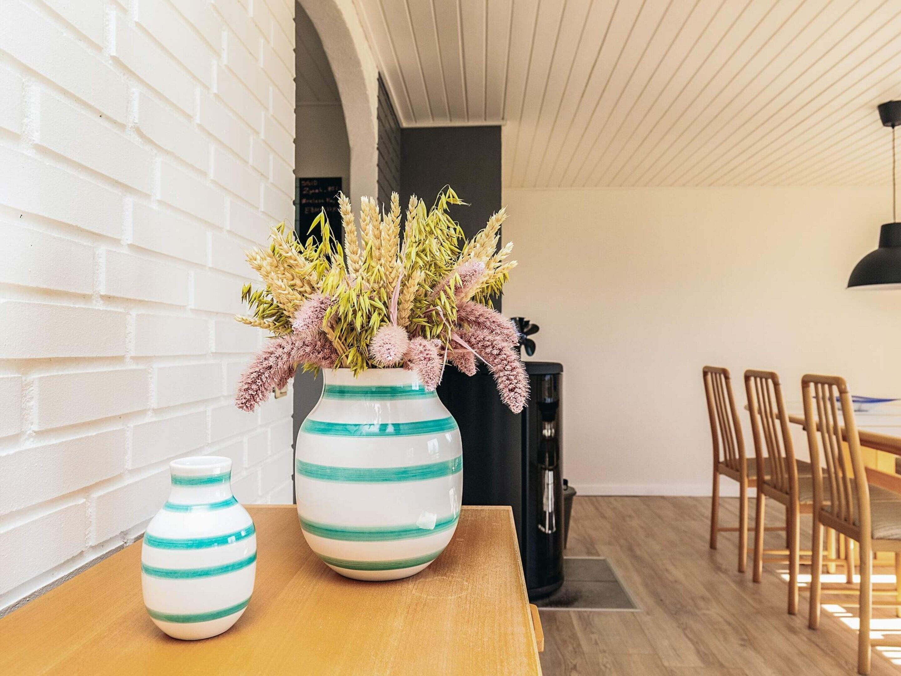 Un comedor con una mesa de madera, dos jarrones a rayas con plantas secas, una cafetera negra y sillas de madera.