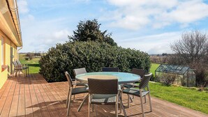 Una terraza de madera con una mesa redonda de vidrio y sillas de mimbre, rodeada de vegetación y un cielo despejado.