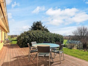 A wooden deck with a round glass table and wicker chairs, surrounded by greenery and a clear sky. - 4 Person Holiday Home in Ostervra (Østervrå)