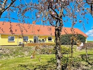 A yellow building with a red roof, a covered patio, and a grassy area with trees. - 4 Person Holiday Home in Ostervra (Østervrå)