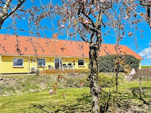 A yellow building with a red roof, a covered patio, and a grassy area with trees. - 4 Person Holiday Home in Ostervra (Østervrå)