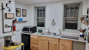 Una cocina con paredes de azulejos blancos, gabinetes de madera, microondas, lavabo y cafetera. Hay cuadros enmarcados en la pared y una estantería con un extintor.