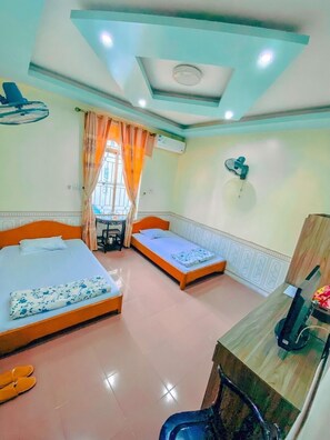 A hotel room with two beds, a desk, a chair, a fan, and a window with curtains. - Pham Gia Hotel Ha Giang - by Bay Luxury (Ha Giang)