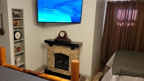 A living room with a stone fireplace, a television mounted on the wall, and a dining area with a table and chairs.