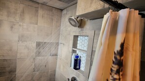 A shower area with tiled walls, a showerhead, a window with a hexagonal pattern, and a shelf with bottles.