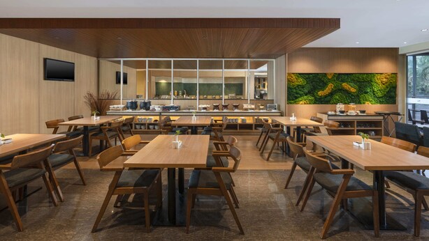 A modern dining area with wooden tables and chairs, a kitchen in the background, and a large green wall art piece. - Swiss-Belexpress Cideng (Jakarta)
