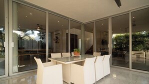 Un espace de repas moderne avec une table en verre et des chaises blanches, de grandes fenêtres et une vue sur l’extérieur.