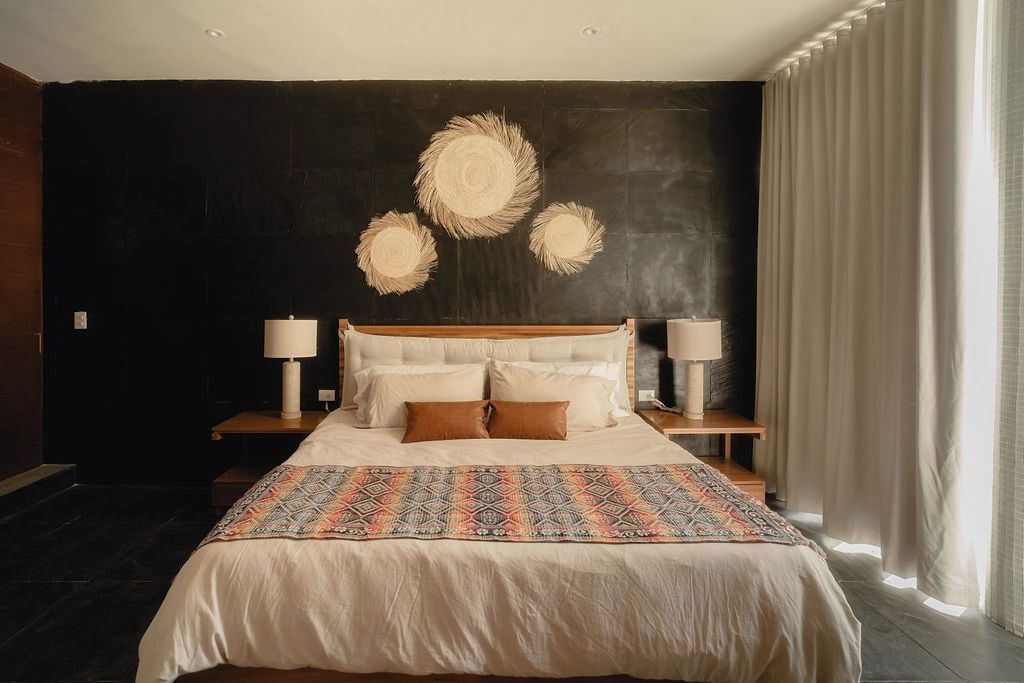 A bedroom with a large bed, two bedside tables with lamps, a dark wall with a circular design, and a window with curtains.