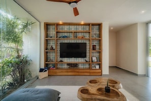 A modern living room with a large flat-screen TV, wooden shelving with decorative items, and a ceiling fan.