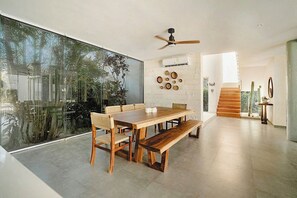 Een ruime eethoek met een grote tafel, banken en stoelen, een plafondventilator en uitzicht door een glazen wand.