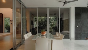 Un espace repas moderne avec une table en verre et des chaises blanches, un ventilateur de plafond et une vue sur la verdure à l’extérieur.