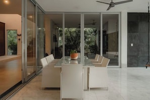 A modern dining area with a glass table and white chairs, a ceiling fan, and a view of greenery outside.