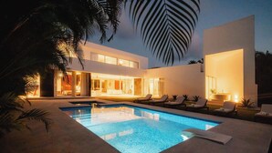 Une maison moderne dotée d’une piscine, de chaises longues et d’un intérieur bien éclairé.