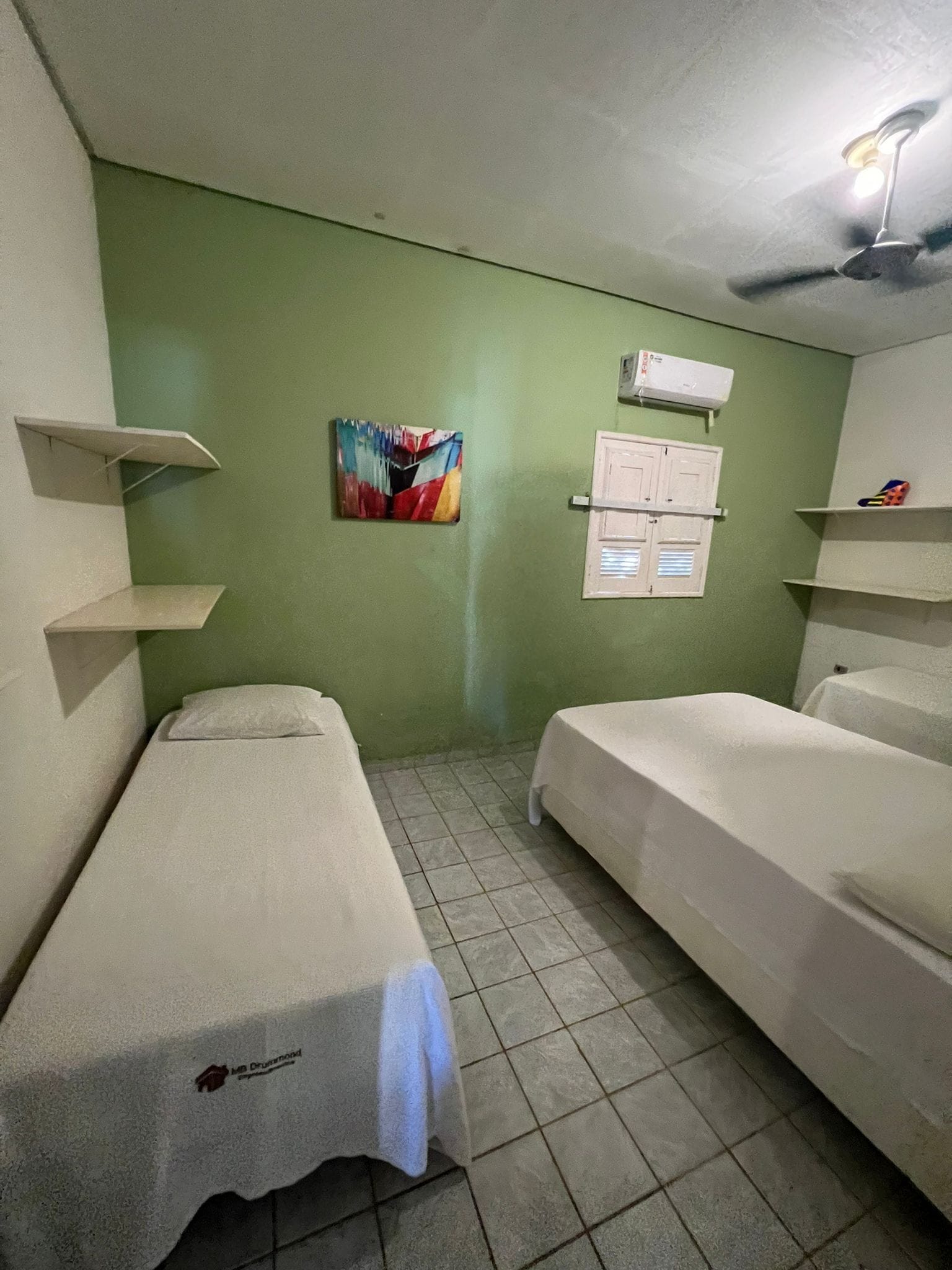 Una habitación pequeña y limpia con dos camas, una pared verde, un cuadro y un ventilador de techo.