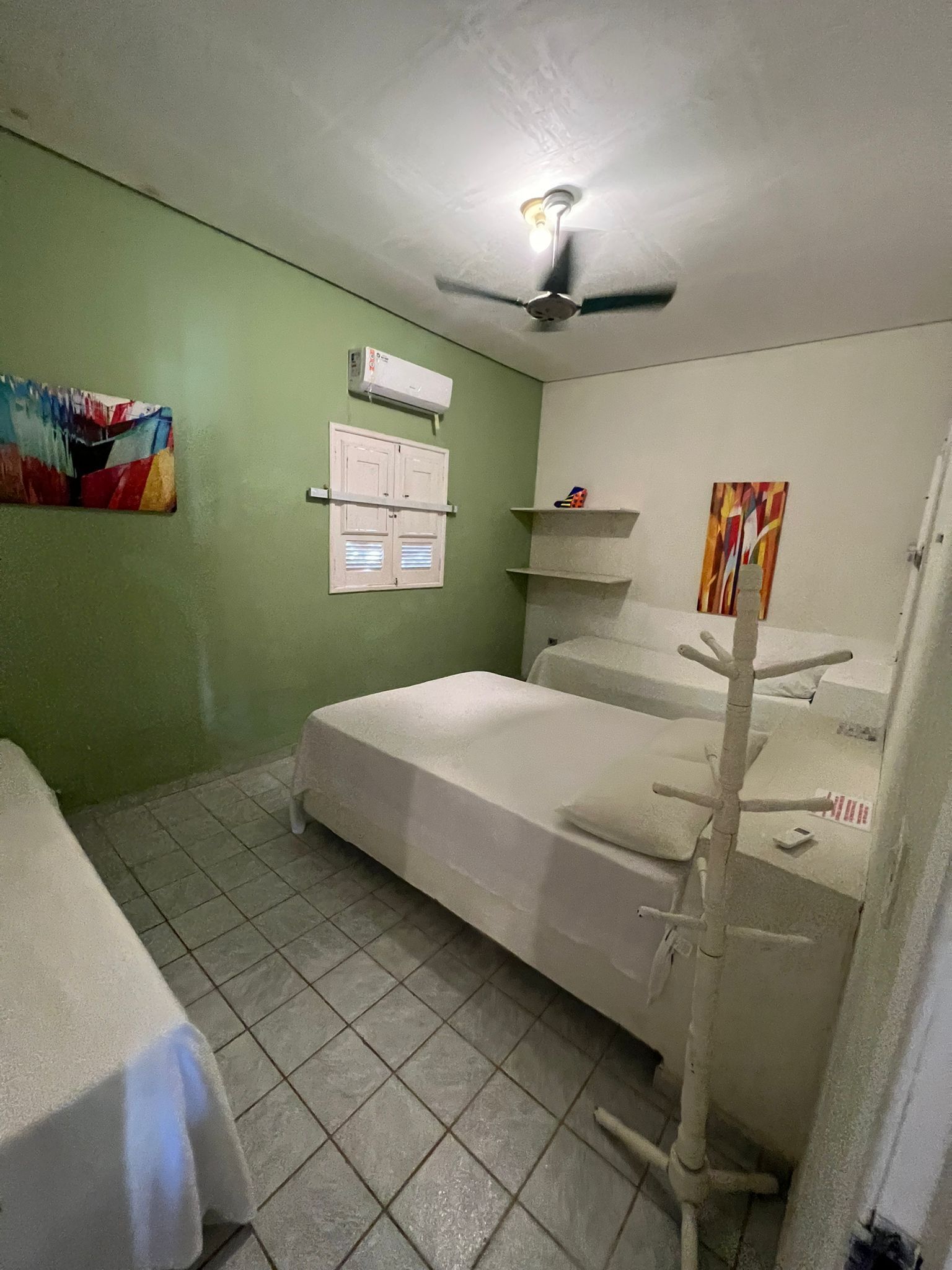 Ein Zimmer mit Bett, Deckenventilator, Fenster mit Jalousien, Ablagefläche und einem Bild an der Wand.