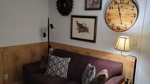 Un salon avec un canapé de couleur violet foncé, deux coussins à motifs, une horloge ronde en bois, un tableau représentant un oiseau encadré, une couronne décorative et une lampe posée sur une table d’appoint en bois.