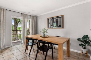 A dining area with a wooden table, black chairs, a potted plant, and a framed artwork on the wall.