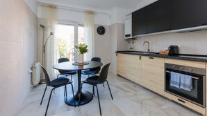 A modern kitchen with a dining area, featuring a round table with chairs, a built-in oven, and a large window with curtains.