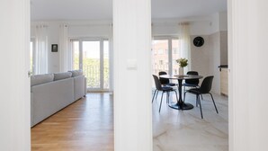 A modern living room with a grey sofa, a dining area with a round table and four chairs, and a large window with a view of the outside.