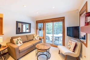 A modern living room with a sofa, a round coffee table, a TV, and a sliding glass door leading to an outdoor area. - Forest Setting, Shared Hot Tub, Ski Home, Parking (Whistler)