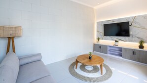 A modern living room with a grey sofa, a round wooden coffee table, a TV mounted on a marble wall, and a built-in kitchen area with grey cabinets.