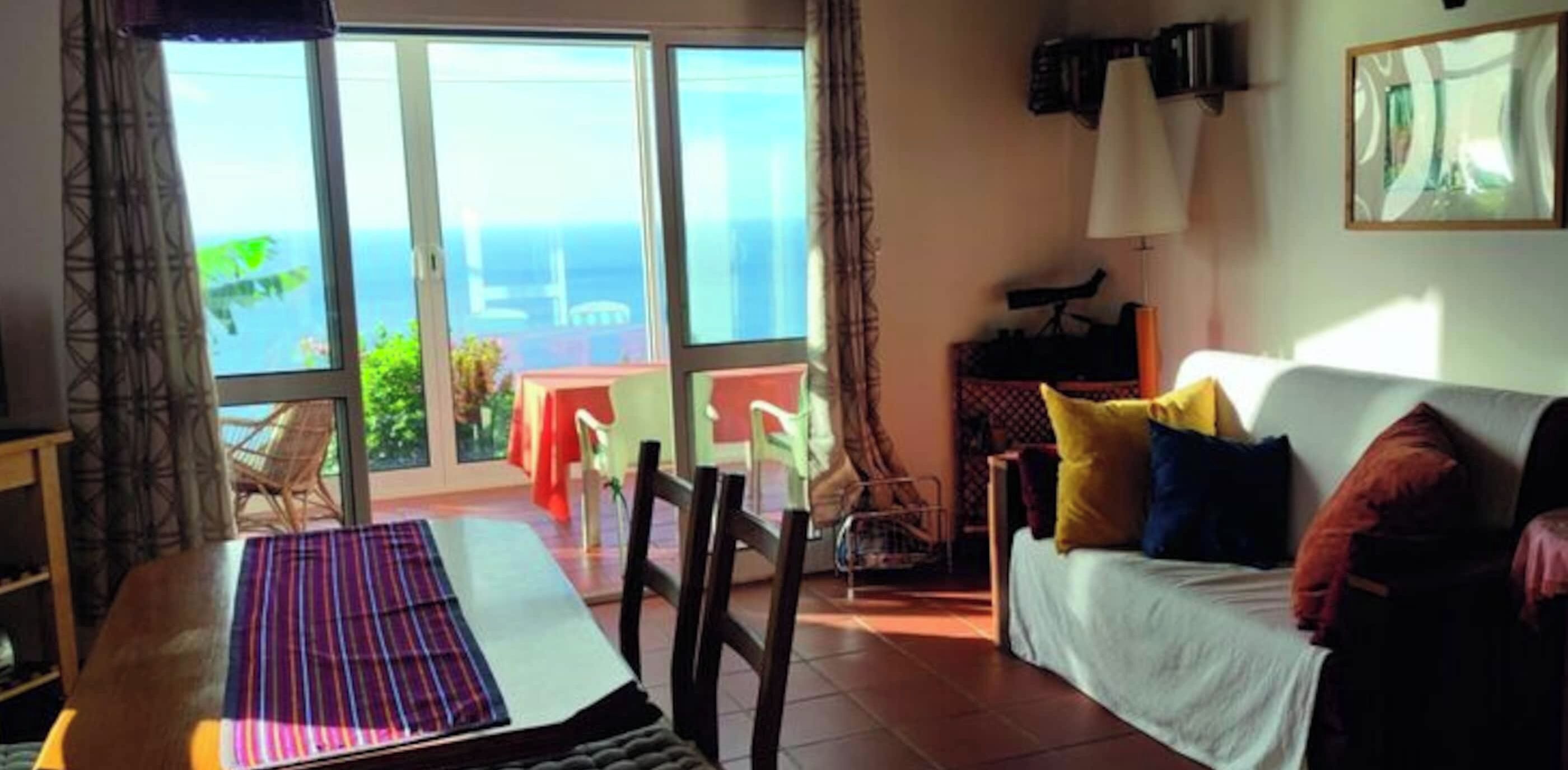 A room with a sofa, a dining table, and a view of the sea through large windows.