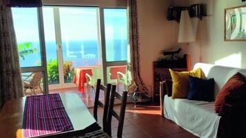 A room with a sofa, a dining table, and a view of the sea through large windows.