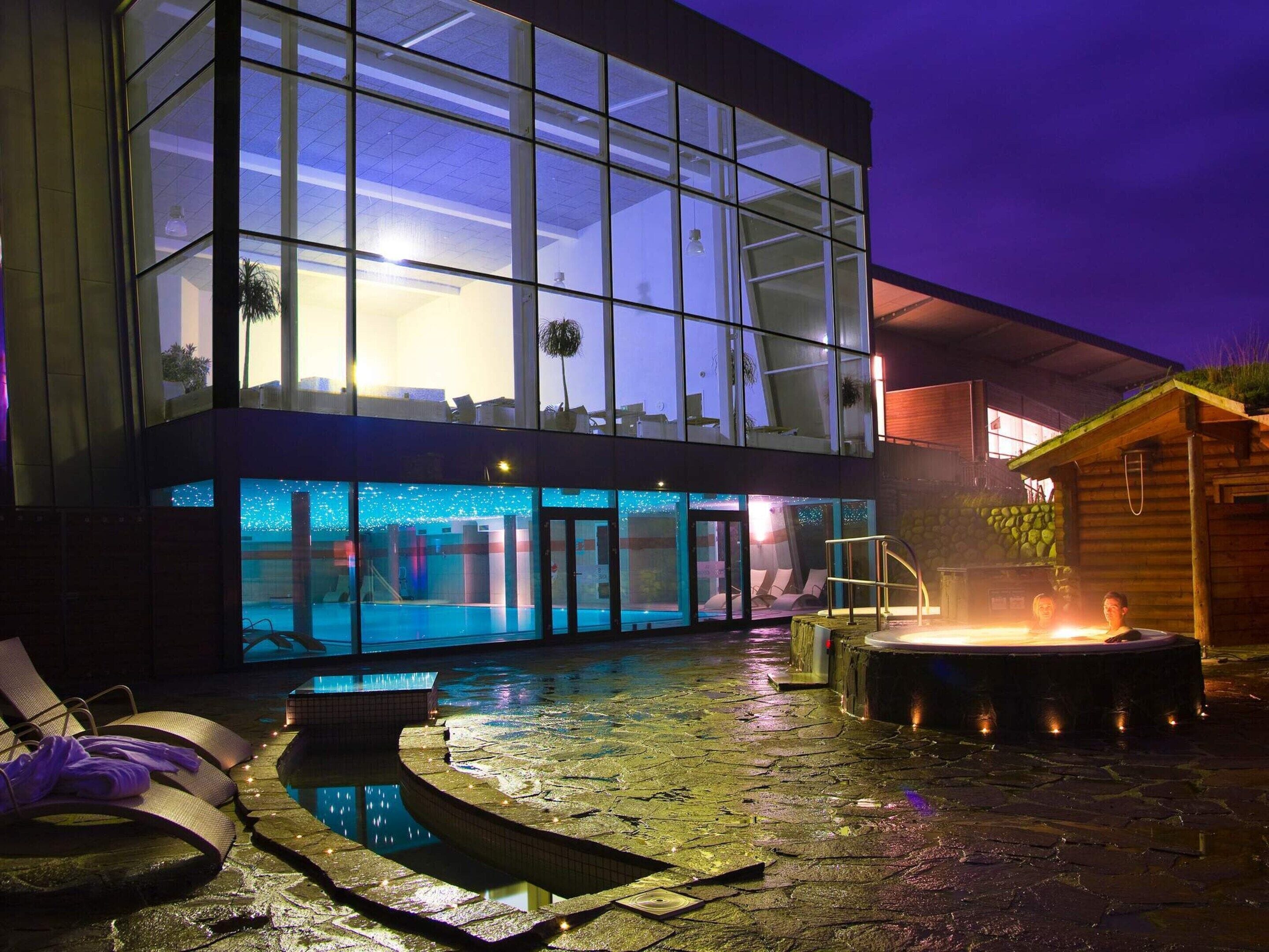 Un bâtiment moderne doté d’une façade en verre, d’une piscine et d’un espace équipé d’un jacuzzi.