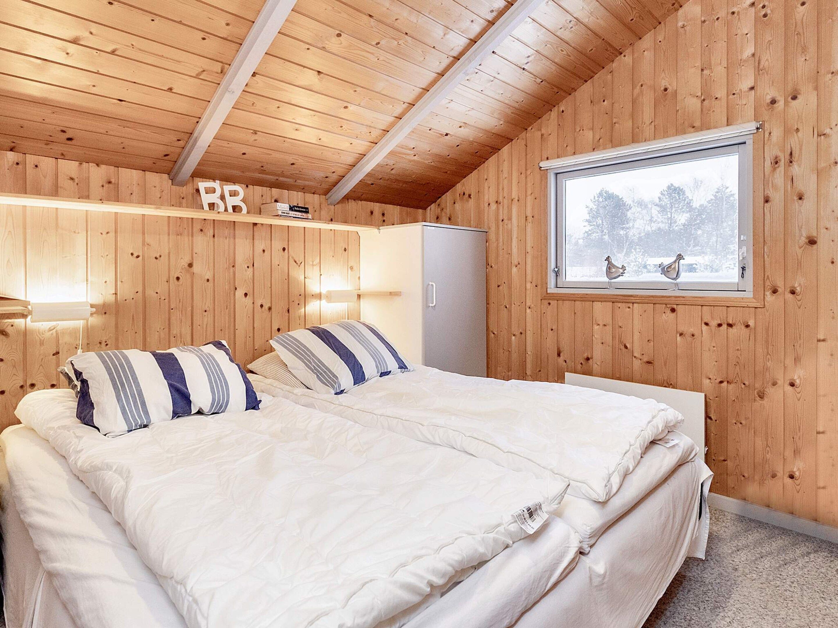 Een kamer met houten lambrisering, een bed, een raam met uitzicht op bomen en een plank met boeken.