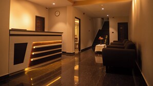 Zona de recepción de un hotel con mostrador, un reloj, un ventilador de techo y una escalera que sube a otro piso.