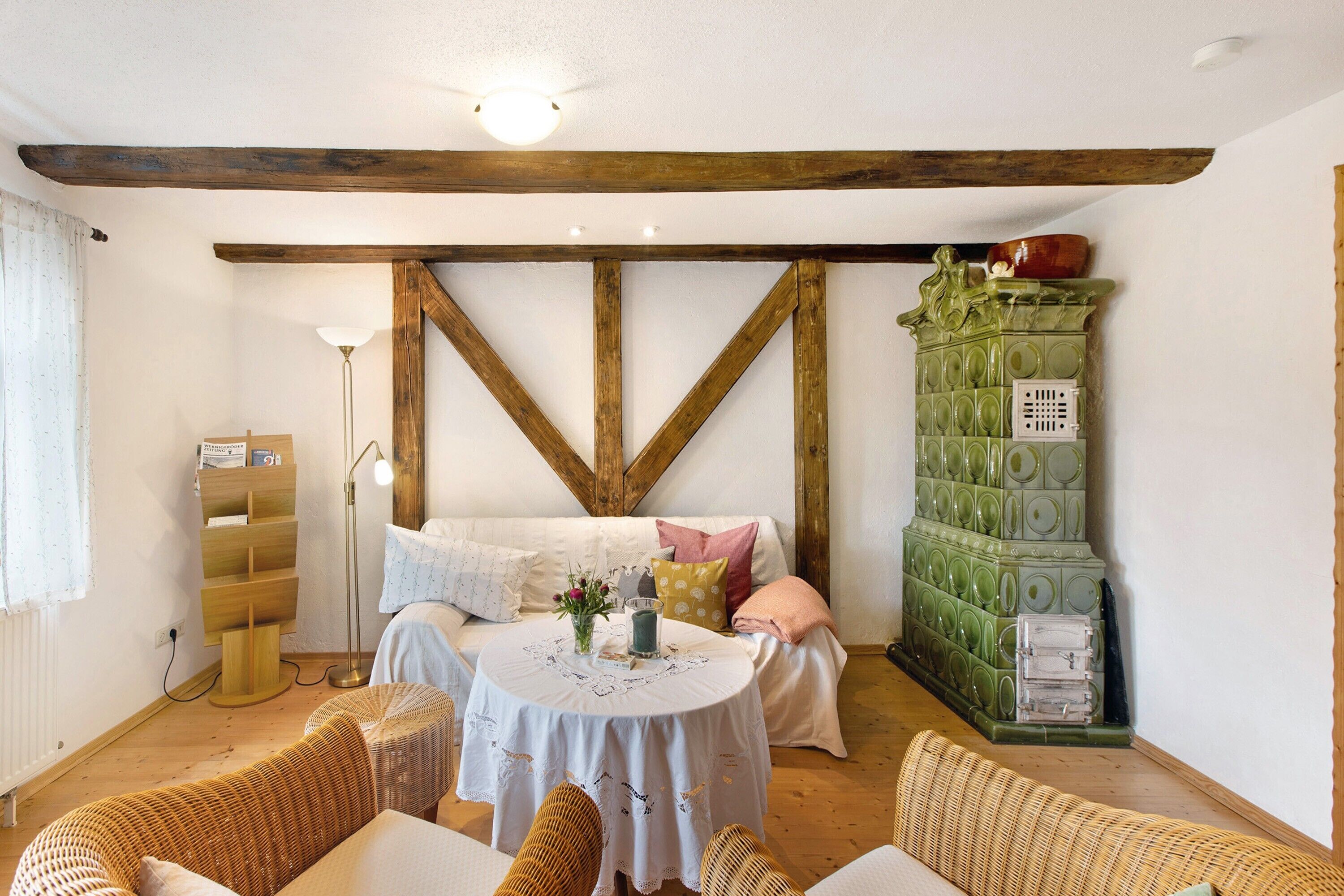 Una habitación acogedora con techo de madera, estufa de azulejos verdes, una mesa redonda con mantel blanco y mobiliario de mimbre.