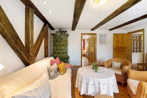 Une pièce chaleureuse avec un plafond en bois, un meuble vert, une table ronde recouverte d’une nappe en dentelle et des meubles en osier.